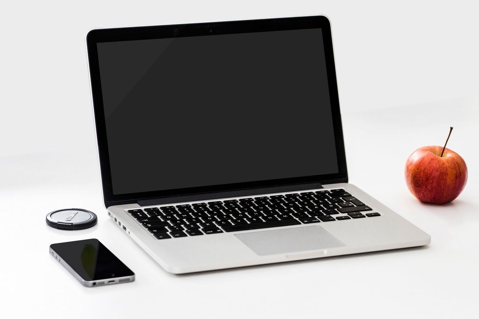 Laptop met donker scherm, smartphone, lensdop en rode appel op wit oppervlak